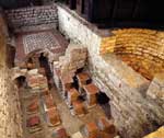 he west wing of Chedworth, showing the mosaic floor of the tepidarium (warm room), wall flues, hypo callor pillars of the caldarium (hot room) and the semi-circular hot bath. 